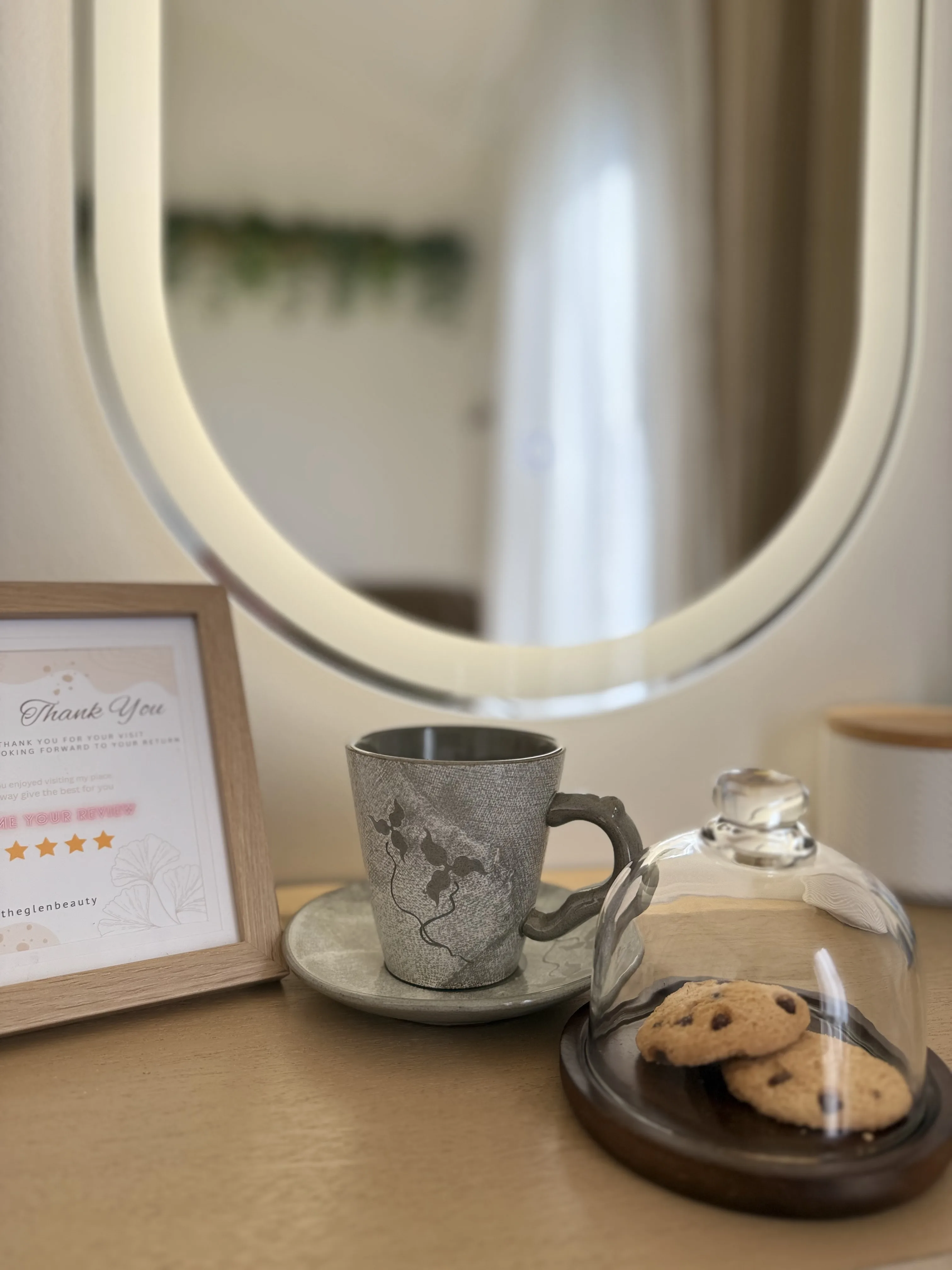 Vanity vignette — LED oval mirror, a handwritten thank-you review card, a textured mug and saucer, glass cloche of cookies