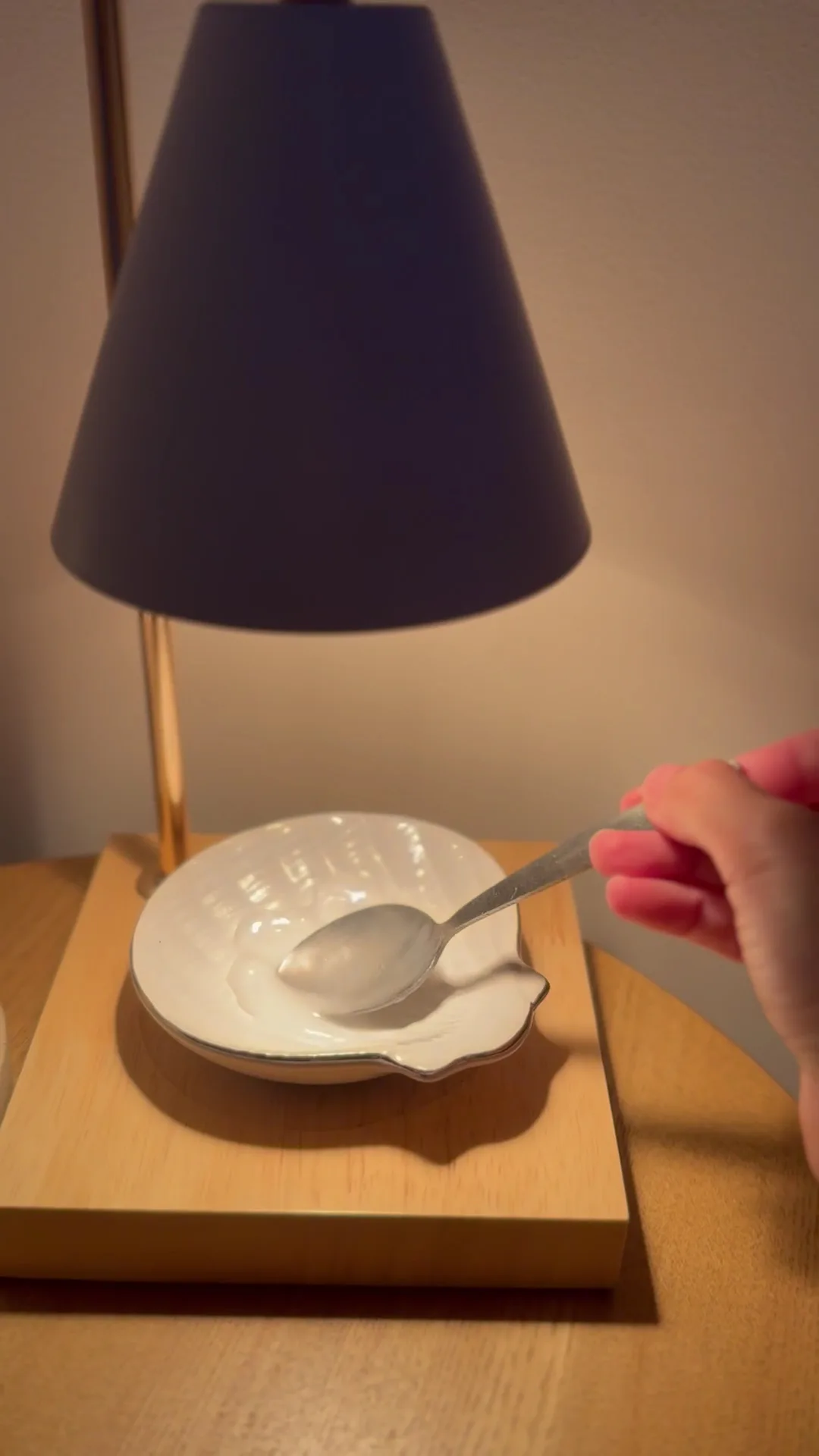 Candle-warmer lamp melting balm in a shell dish, a hand lifting a spoon — a soft ambient detail