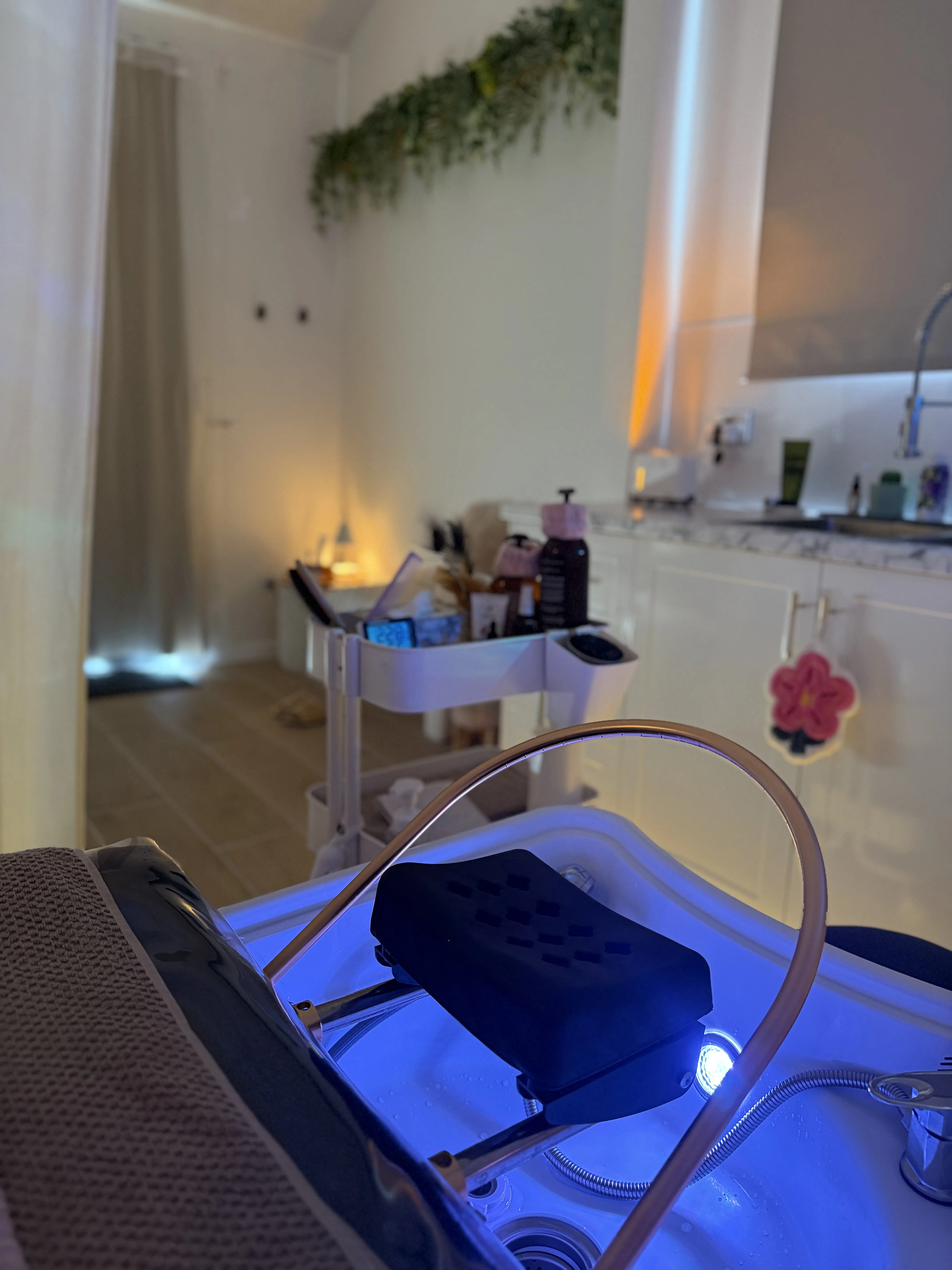 The treatment room looking down its full length — treatment bed, product trolley, blue-lit basin and marble kitchenette at the back