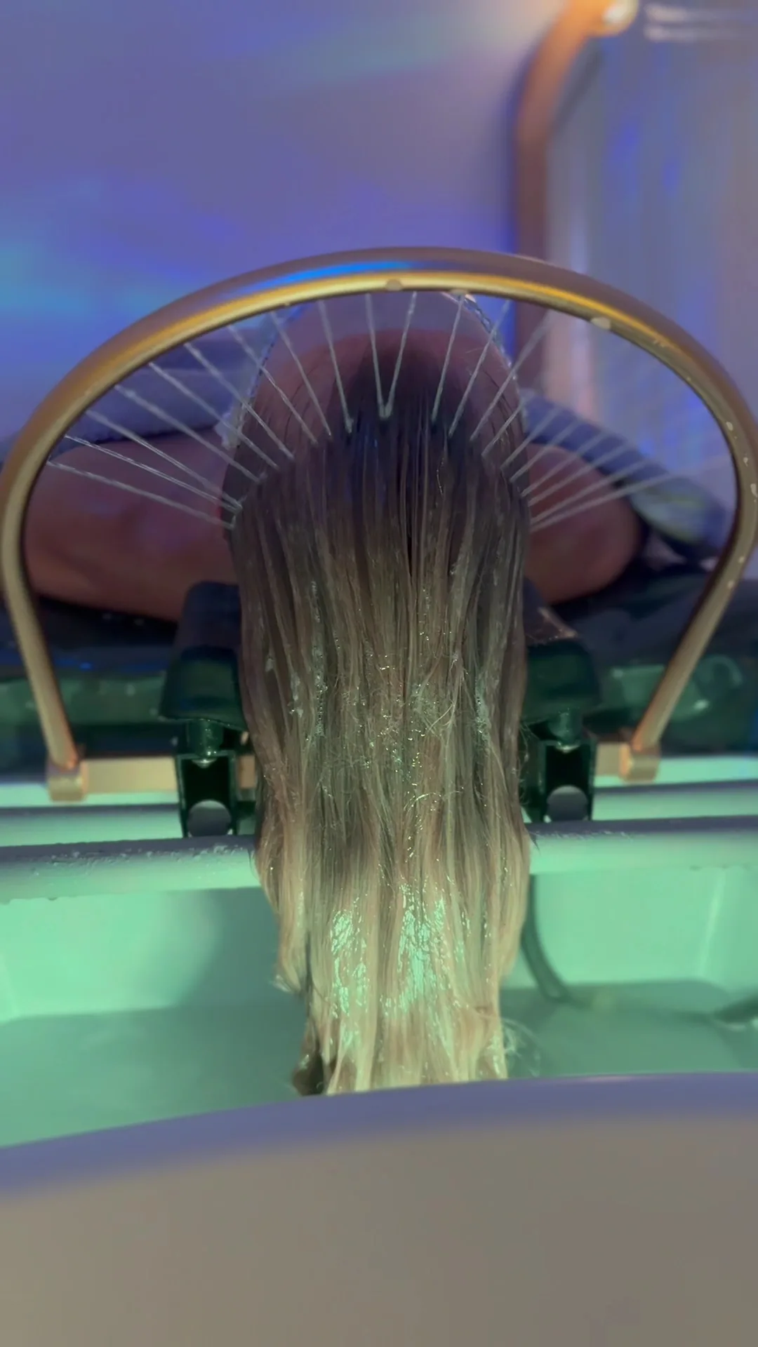 Water halo rinsing blonde-ombre hair over the gold ring under blue-green basin light